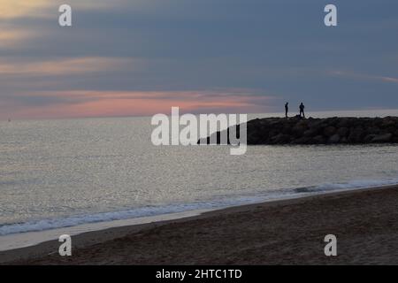 Zwei Menschen stehen auf den Felsen vor einem ruhigen Meer, das von einem Sandstrand erfasst wurde Stockfoto