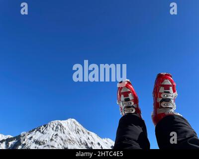 Frauenbeine in roten Skischuhen ruhen nach einer Skitour am sonnigen Tag, verschneite Alpen im Hintergrund. Hochwertige Fotos Stockfoto