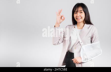 Arm gebrochen. Geschäftsfrau zuversichtlich Lächeln gebrochenen Arm nach Unfall tragen Armschiene für Behandlung zeigen OK Zeichen, glücklich asiatische weibliche Sling Stützhand Stockfoto