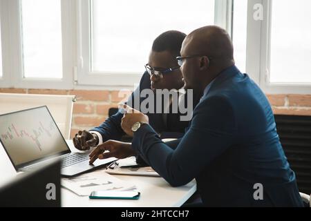 Zwei dunkelhäutige Partner eines afroamerikanischen Geschäftsmannes arbeiten bei einem Meeting auf einem Laptop im Büro. Erschwingliche private Kurse und Entfernung Stockfoto