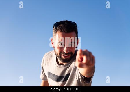 Bärtiger Mann, der mit dem Finger auf die Kamera zeigt, mit blauem Himmel im Hintergrund, Platz für Text kopieren Stockfoto