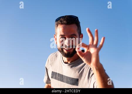 Bärtiger lächelnder Mann, der die Kamera anschaut und das OK-Symbol mit seiner Hand mit dem blauen Himmel im Hintergrund macht, Platz für Text kopieren Stockfoto