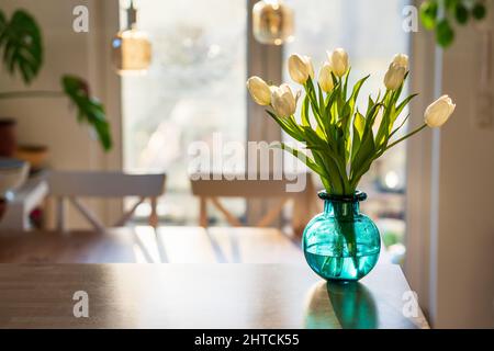 Ein Bouquet aus weißen Tulpen in einer blau-grünen Glasvase in der hellen Morgensonne. Modernes Innendesign mit grünen Pflanzen und frischen Frühlingsblumen in allen r Stockfoto