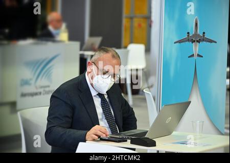 Der Geschäftsmann, der auf der internationalen Messe am Schreibtisch arbeitet, trägt eine obligatorische Gesichtsmaske Stockfoto