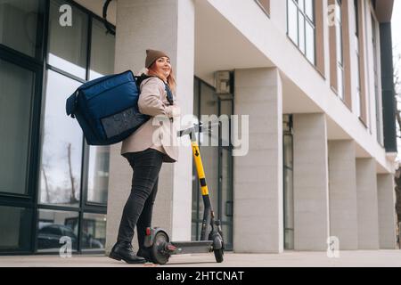 Low-Angle-Ansicht von positiven weiblichen Kurier mit großen Thermo-Rucksack stehen mit Elektroroller in der Stadt Straße Blick auf die Kamera. Stockfoto