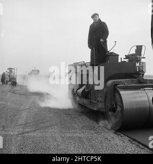 RAF Gaydon, Gaydon, Stratford-on-Avon, Warwickshire, 21/01/1953. Ein Mann, der während der Asphaltverlegung auf einer neuen Start- und Landebahn im Bau auf dem Flugplatz Gaydon auf einer Walze steht. Anfang 1952 begannen die Arbeiten am Bau einer Start- und Landebahn auf dem Flugplatz Gaydon. Im Rahmen des Projekts wurde eine ehemalige Start- und Landebahn, die nach dem Zweiten Weltkrieg weitgehend aufgegeben wurde, durch eine fast 1 3/4 Meilen lange und 200 Fuß breite, neu errichtete Start- und Landebahn ersetzt. Ebenfalls gebaut wurden Zufahrtswege und ein Taxi-Gleis. Die Start- und Landebahn wurde auf 8 Zoll Hardcore gebaut, wobei vier Zoll Beton darauf gelegt wurden, gefolgt von 12 Zoll H Stockfoto