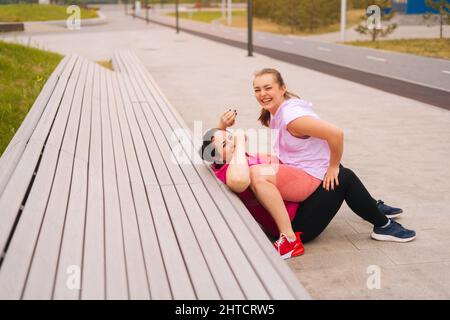 Lachende, fette junge Frau trainiert mit einem fröhlichen Fitnesstrainer auf einer Bank im Freiluftgymnastik im Stadtpark. Stockfoto