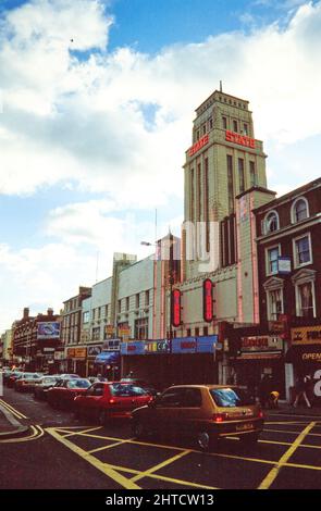 Mecca Bingo Club, Kilburn High Road, Kilburn, Brent, London, 1990-2004. Der Mekka Bingo Club und die Kilburn High Road von Norden aus gesehen. Das Gaumont State Theatre wurde 1937 eröffnet. Es war eines der größten Kinos in Europa und das größte in England mit einer Kapazität von 4.004 Sitzplätzen. Das äußere Design ist angeblich vom Empire State Building in New York inspiriert. Das Gebäude wurde später in eine Tanzhalle, ein Kino und eine Bingohalle unterteilt, bevor es ab etwa 1990 ausschließlich als Bingohalle genutzt wurde. 2010 wurde das Gebäude als Kirche wiedereröffnet. Stockfoto