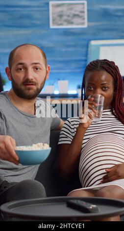 POV der interracial Familie Blick auf Kamera auf der Couch sitzen. Gemischtes Rennpaar erwartet Kind beim Fernsehen. Kaukasischer Mann bringt Glas Wasser für schwarze Schwangere Stockfoto