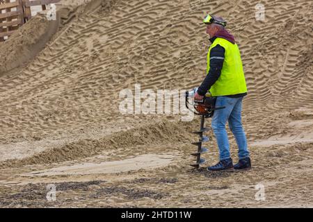 Mann in hoher Weste bohrt Löcher für Stellen in den Vorbereitungen natürlich bei Motocross-Rennen am Weymouth Beach, Weymouth, Dorset UK im Oktober Stockfoto