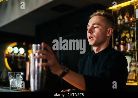 Nahaufnahme eines fokussierten Barkeepers, der einen erfrischenden alkoholischen Cocktail zubereitet und hinter der Theke in einem modernen dunklen Nachtklub steht. Stockfoto