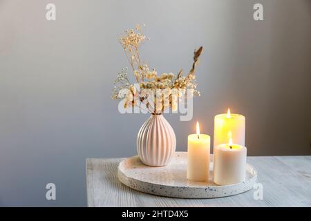 Ein Bouquet aus getrocknetem Gypsophila und Eukalyptus in einer kleinen weißen gewellten Vase und drei große brennende Kerzen auf einem runden Tablett befinden sich auf einem beigen Tisch. Pl Stockfoto