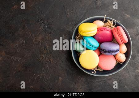 Bunte Makronen und Schokoladeneier in Keramikschale auf schwarzem Beton-Hintergrund. Draufsicht, Kopierraum, flach liegend, Stillleben. Frühstück, morgens Stockfoto