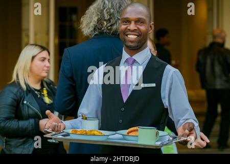 Lächelnder schwarzer Caterer hält ein Tablett bei einem Ereignis auf einem überfüllten Hintergrund Stockfoto