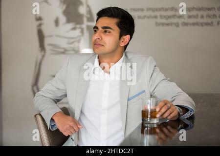 Nahaufnahme des südasiatischen Unternehmers, der auf dem grauen Hintergrund sitzt und trinkt Stockfoto