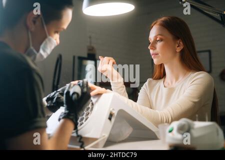 Seitenansicht einer attraktiven jungen Frau, die behandelte Nägel untersucht und die Arbeit eines Manikuristen im Kosmetiksalon bewertet. Stockfoto