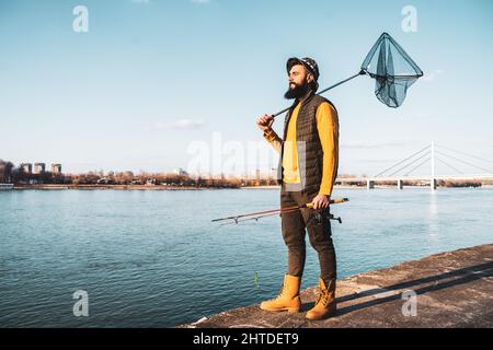 Bild eines Fischers, der Angelrute und Fischernetz hält, während er an einem Fluss steht. Stockfoto