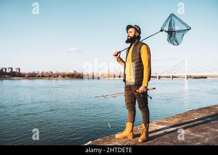 Bild eines Fischers, der Angelrute und Fischernetz hält, während er an einem Fluss steht. Stockfoto