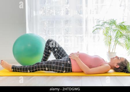 Junge Schwangere, die auf einer Yogamatte liegt, macht Übungen und dehnt sich aus, um während der Schwangerschaft beweglich und aktiv zu bleiben Stockfoto