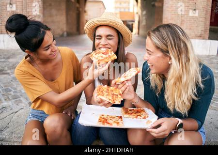 Glückliche Freundinnen essen Pizza Street Food in der Stadt. Weibliche Touristengruppe aus drei Frauen, die Spaß haben. Ein Mädchen füttert das andere. Lifestyle und Stockfoto