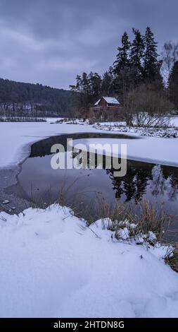Ein vertikaler Blick auf den Fluss, der im Winter durch den schneebedeckten Boden zu einer Hütte gegen Bäume fließt Stockfoto