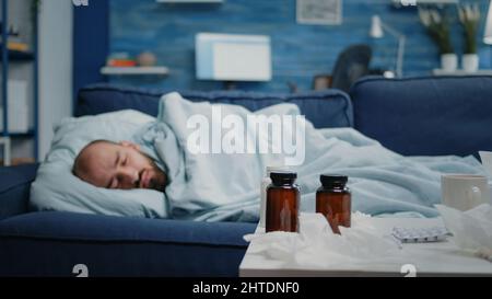 Kranker Mann schläft auf der Couch mit Decke, während er Erkältung und Grippe hat. Person, die wegen einer Viruserkrankung ruht und Flaschen mit Pillen, Medikamenten, einer Tasse Tee und Taschentüchern auf dem Tisch hat. Stockfoto
