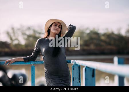 Brünette Frau mit geschlossenen Augen und Hut entspannen auf einem Pier am Fluss. Konzept der Trennung in der Natur. Stockfoto