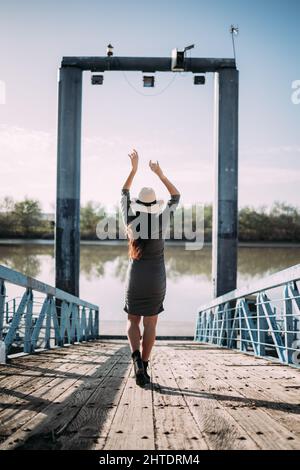 Brünette Frau mit Hut und Arme angehoben entspannend auf einem Pier am Fluss. Konzept der Trennung in der Natur. Stockfoto