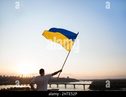 Die ukrainische Flagge flattert im Wind gegen den Sonnenuntergangshimmel. Ein Mann, der auf dem Dach eines Hauses in Kiew steht, hält eine große Flagge. Nationales Symbol von Freed Stockfoto