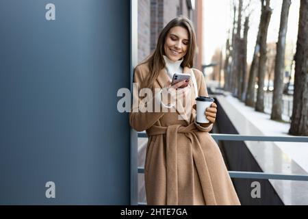 Hübsche Brünette Mädchen in braunem Mantel mit Kaffeetasse mit Smartphone im Freien Stockfoto