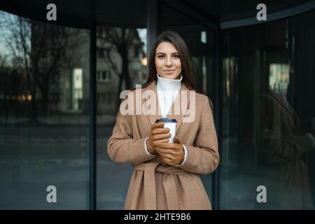 Frau in braunem Mantel mit einer Tasse Kaffee auf dem Hintergrund der Glastüren des Bürogebäudes Stockfoto