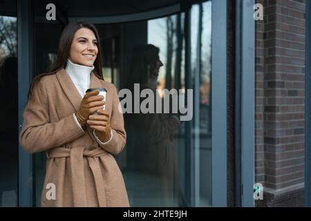 Schöne Geschäftsfrau in elegantem Mantel mit einer Tasse Kaffee steht in der Nähe des Eingangs zum Bürogebäude Stockfoto