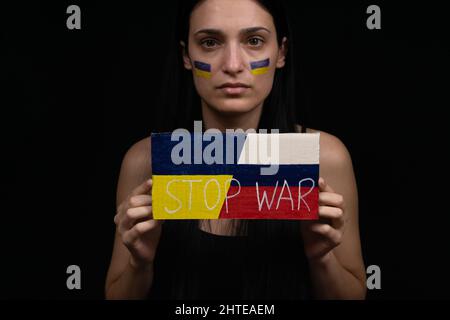 Die ukrainische Frau hält die Inschrift STOP war in ihren Händen. Schwarzer Hintergrund. Auf ihrem Gesicht ist eine gelbe und blaue Flagge gemalt Stockfoto