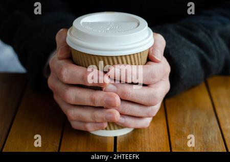 Frauenhände halten Karton umweltfreundliche Einweg-Tasse mit Kunststoffdeckel auf Holztisch mit selektivem Fokus Stockfoto