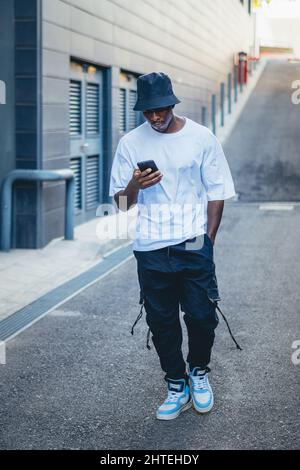 Vertikale Aufnahme eines lässigen jungen schwarzen Models mit einem blauen Hut, der sein Telefon im Freien überprüft Stockfoto