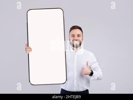 Junger Fröhlicher Mann Hält Smartphone Mit Leerem Bildschirm Und Zeigt Daumen Nach Oben Stockfoto