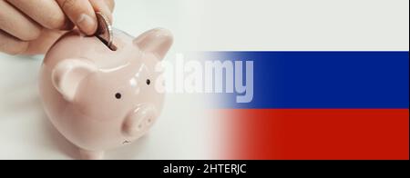 Nahaufnahme einer Frau, die vor dem Hintergrund der russischen Nationalflagge eine Münze in das Sparschwein steckt. Sparkonzept Stockfoto