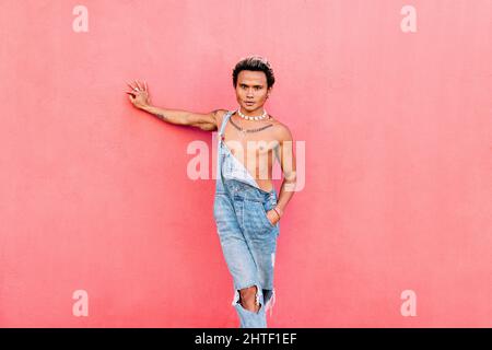 Ein junger, hübscher Mann, der Jeans trägt und an der rosa Wand angelehnt ist und die Kamera anschaut Stockfoto