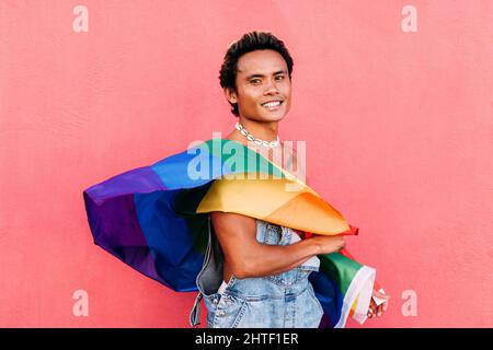 Porträt eines jungen lächelnden Mannes, der draußen mit LGBT-Flagge gegen eine pinke Wand posiert Stockfoto
