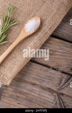 Nahaufnahme von weißem Steinsalz in Holzlöffel von Rosmarin auf Jutestoff am Tisch Stockfoto