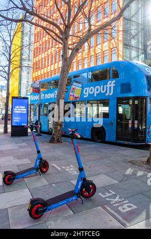Blue Rental Elektro-Scooter parkten auf einem Bürgersteig neben einem blauen Londoner Bus vor modernen Bürogebäuden im Central Saint Giles in London Stockfoto