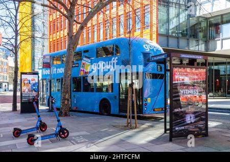 Blue Rental Elektro-Scooter parkten auf einem Bürgersteig neben einem blauen Londoner Bus vor modernen Bürogebäuden im Central Saint Giles in London Stockfoto