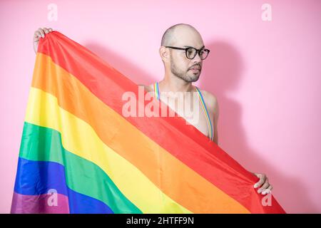 Junge mexikanische Glatze trägt Schwulenhosenträger mit LGBT-Flagge vor einem rosa Hintergrund Stockfoto