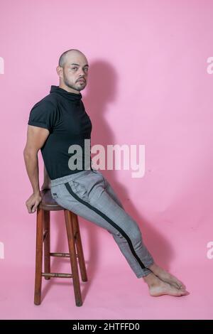 Ganzkörperaufnahme eines jungen, gutaussehenden, kahlen mexikanischen Mannes, der auf einem Stuhl vor einem rosa Hintergrund sitzt Stockfoto