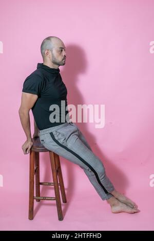 Ganzkörperaufnahme eines jungen, gutaussehenden, kahlen mexikanischen Mannes, der auf einem Stuhl vor einem rosa Hintergrund sitzt Stockfoto