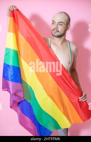 Junge mexikanische Glatze trägt Schwulenhosenträger mit LGBT-Flagge vor einem rosa Hintergrund Stockfoto