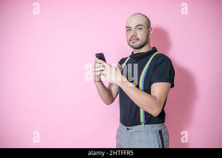 Junge mexikanische Glatze Mann trägt Homosexuell Stolz Hosenträger posiert mit einem Telefon vor einem rosa Hintergrund Stockfoto