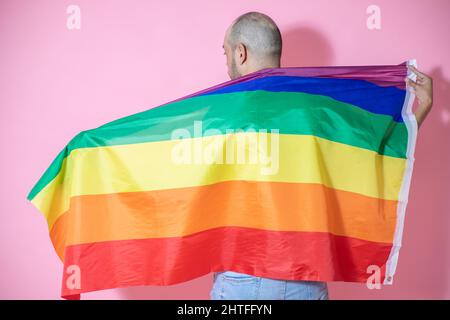 Rückansicht eines jungen, hübschen, kahlen Mannes aus Mexiko, der mit einer LGBT-Flagge vor einem rosa Hintergrund posiert Stockfoto