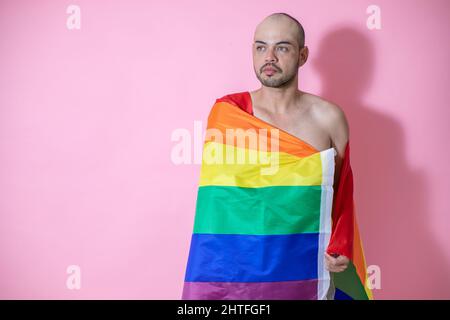 Porträt eines jungen, gutaussehenden mexikanischen Glatzkopfes, der mit LGBT-Flagge vor einem rosa Hintergrund posiert Stockfoto