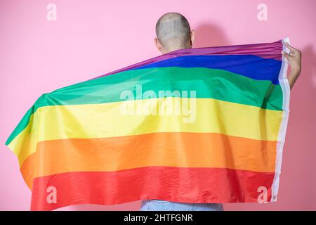 Rückansicht eines jungen, hübschen, kahlen Mannes aus Mexiko, der mit einer LGBT-Flagge vor einem rosa Hintergrund posiert Stockfoto